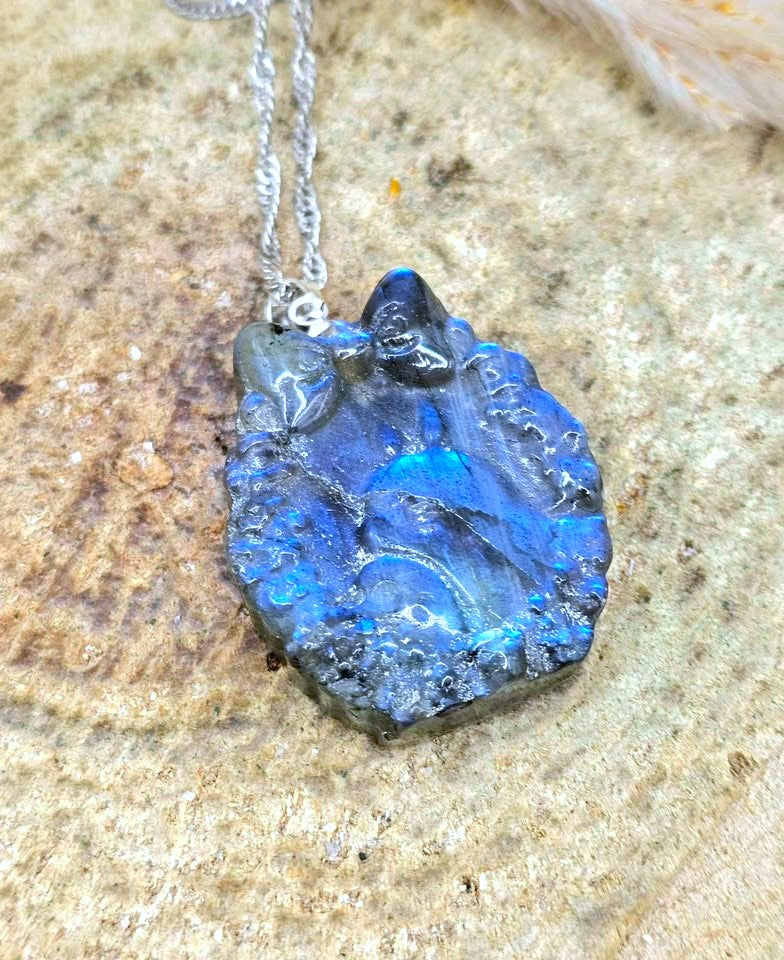 A raw blue labradorite crystal pendant necklace on a silver chain, displayed on a textured beige stone surface.
