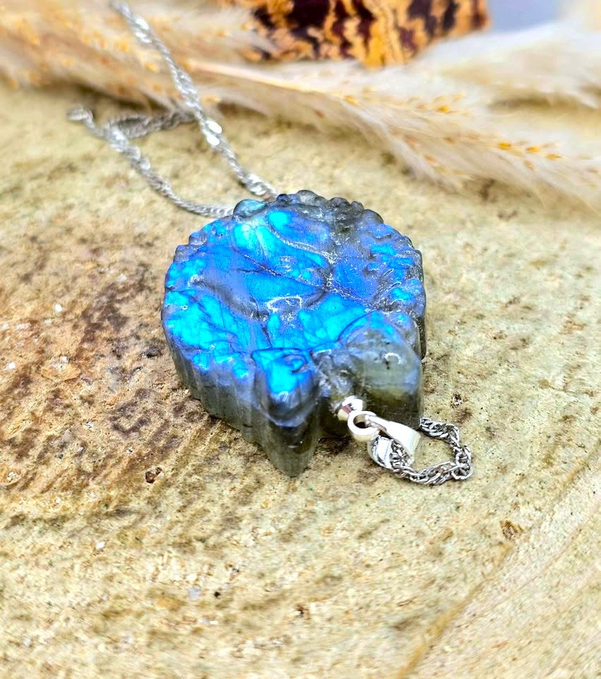A silver necklace featuring a polished labradorite pendant with vibrant blue and green iridescent flashes, resting on a textured stone surface.