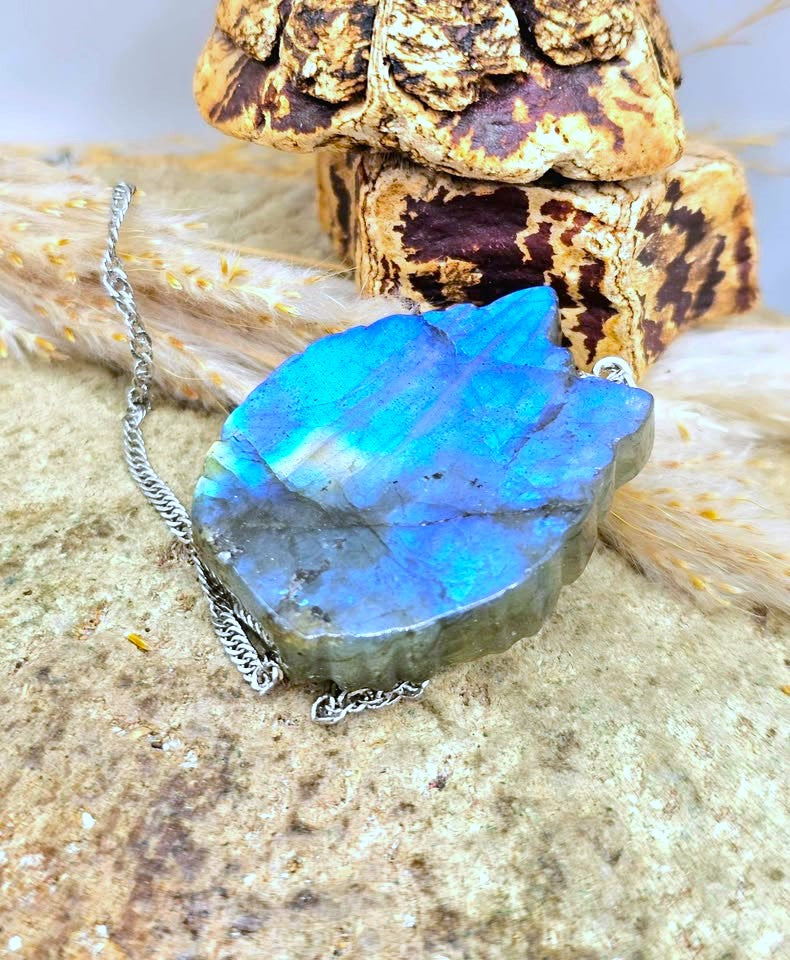A raw labradorite gemstone pendant necklace with a silver chain, displayed on a textured stone surface with dried grass and a decorative block in the background.