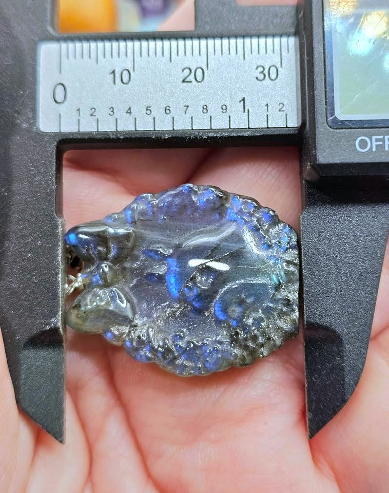 A polished blue-gray labradorite gemstone held in a hand under a digital caliper to show its size. The gemstone displays a sparkling blue and gray play-of-color pattern.