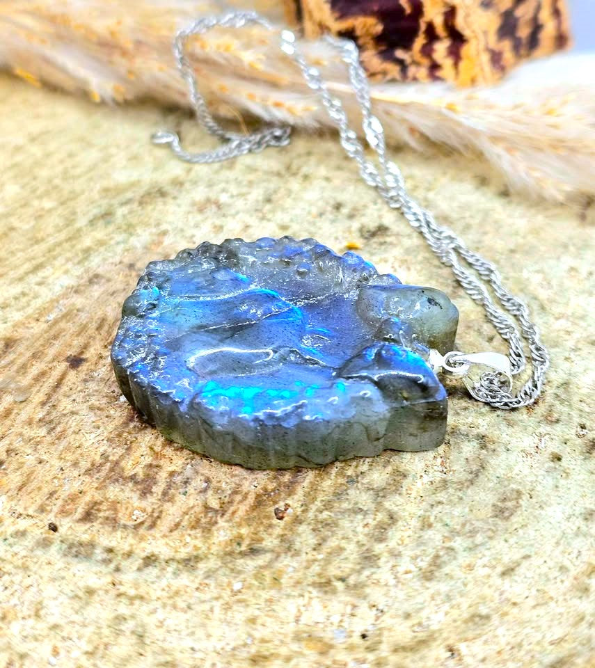 A raw labradorite stone pendant necklace with a silver chain, displayed on a textured beige stone surface.