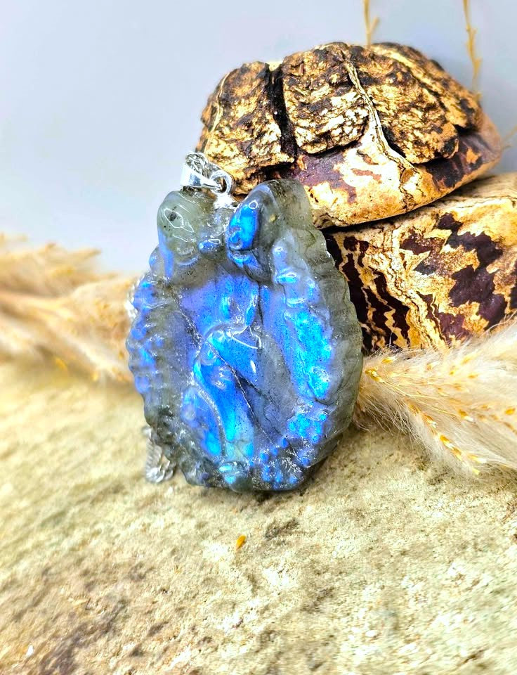 A blue iridescent labradorite pendant with a silver bail, displayed on a textured stone surface with dried grass and a beetle shell in the background.