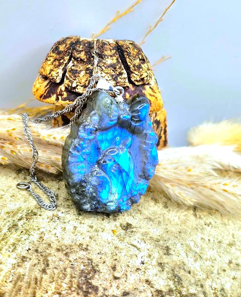 A raw, polished blue opal pendant necklace with a silver chain, displayed on a textured stone surface with dried grass and a gold-toned decorative piece in the background.