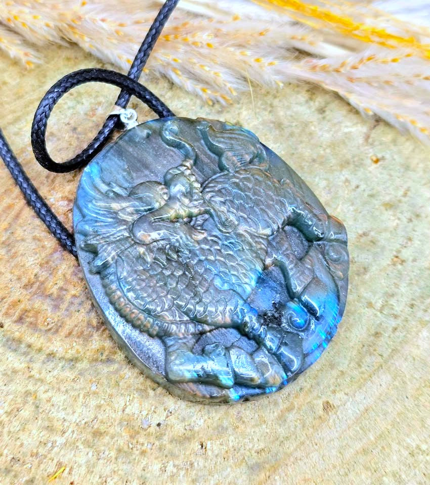 A carved labradorite pendant necklace featuring a detailed dragon design, suspended from a black braided cord and displayed on a textured wood slice background.