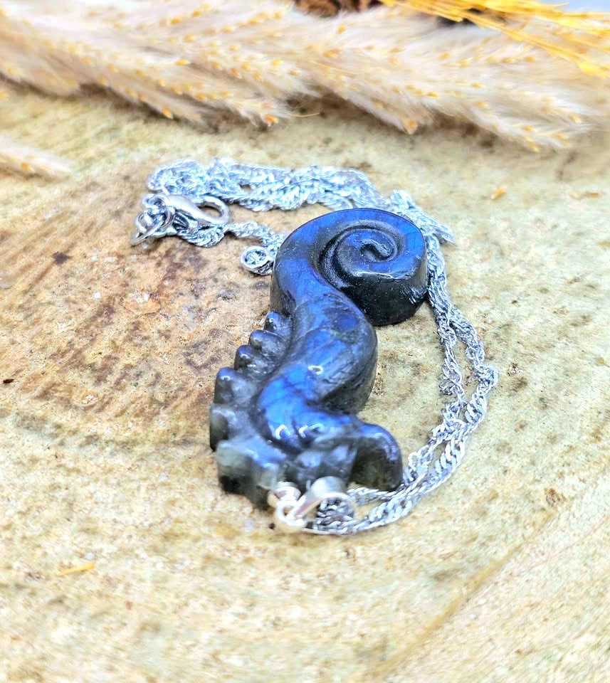 A dark blue carved pendant shaped like a spiral shell or snail, attached to a silver chain, displayed on a textured beige stone surface with dried grass in the background.