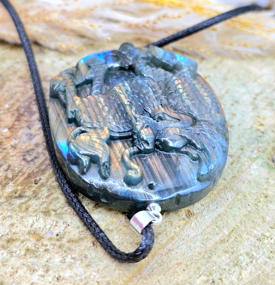 A carved oval labradorite pendant necklace featuring a detailed scene of figures and animals, resting on a textured stone surface. The pendant has a black cord and a silver clasp.