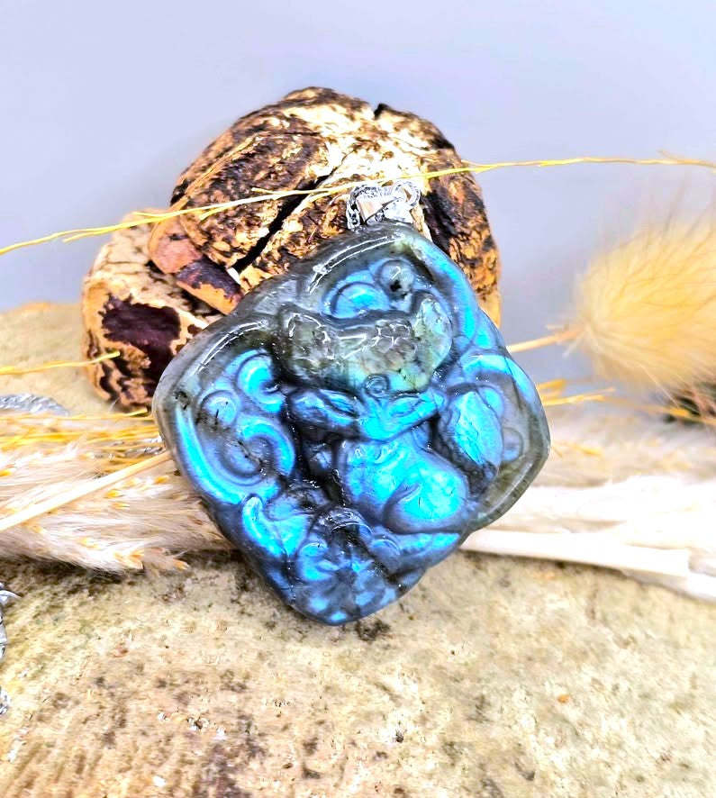 A carved blue labradorite pendant featuring a stylized animal design, displayed on a textured stone surface with dried grass and a decorative rock in the background.