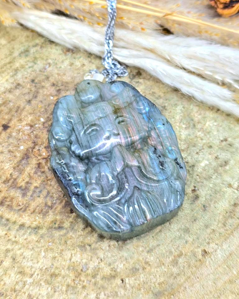 A carved labradorite pendant featuring a stylized face, suspended from a silver chain, resting on a textured stone surface.