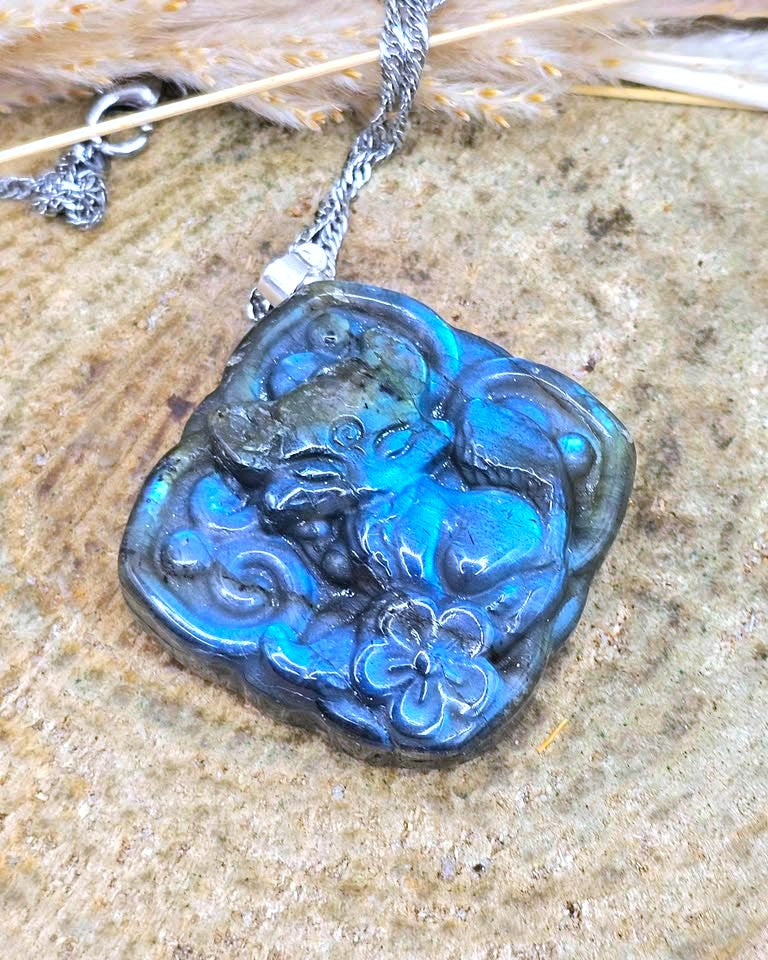 A close-up of a carved labradorite pendant necklace featuring a fox and flower motif, resting on a textured stone surface with dried grass.