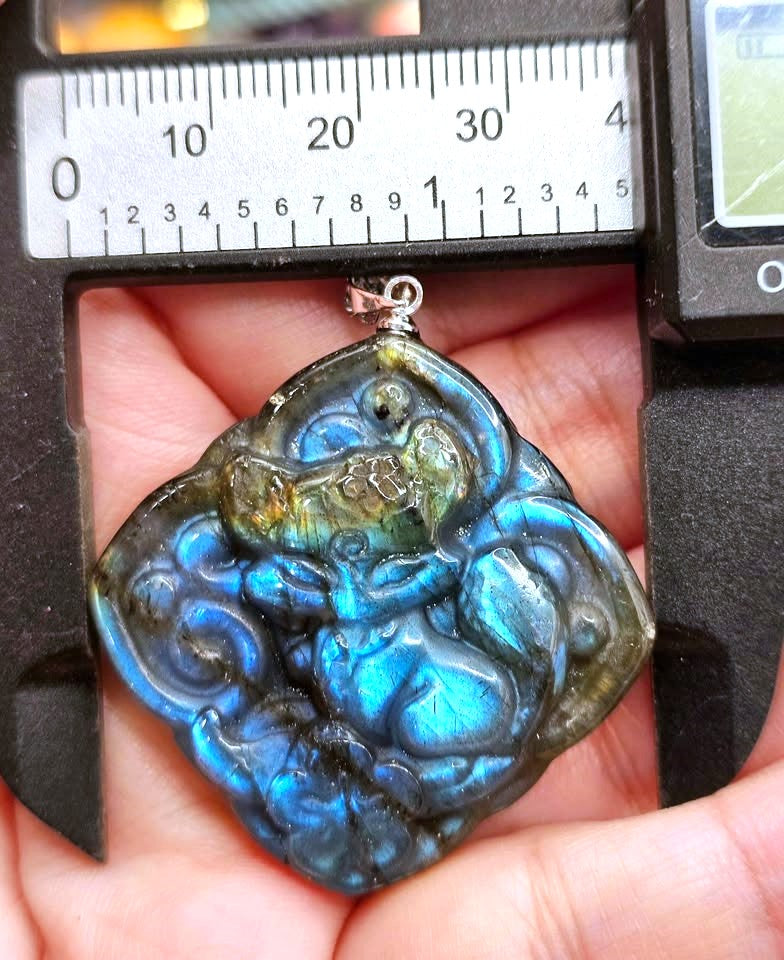 A close-up of a carved labradorite pendant held in a hand under a measuring caliper. The pendant features a detailed, stylized face with a shimmering blue, green, and brown iridescent labradorite finish.