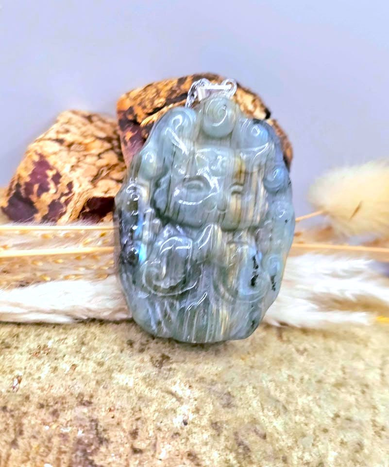 A carved jadeite pendant with a blue-green iridescent sheen, featuring a seated figure design, displayed on a textured stone surface with dried grass and a shell.