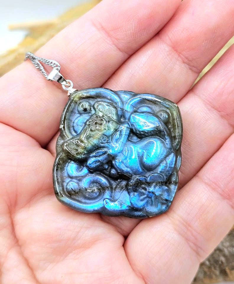 A close-up of a hand holding a square pendant made of iridescent labradorite stone with a silver bail and chain.