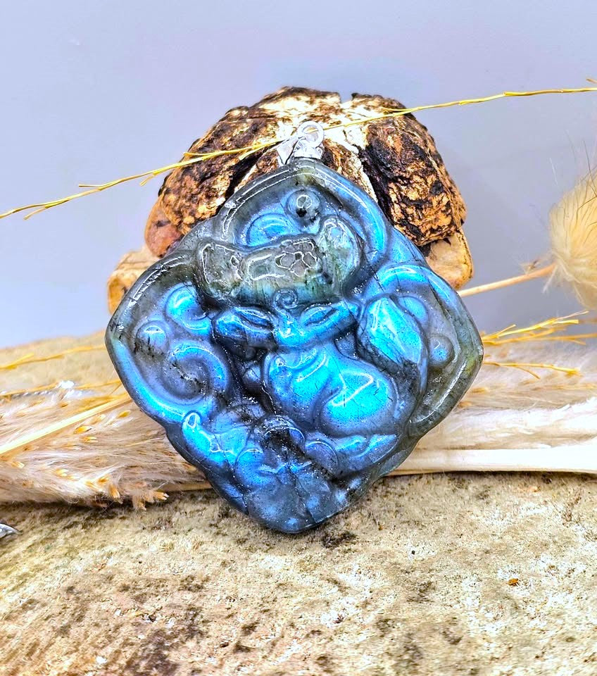 A carved blue labradorite pendant featuring a stylized monkey figure, displayed on a textured stone surface with dried grass and a decorative shell.