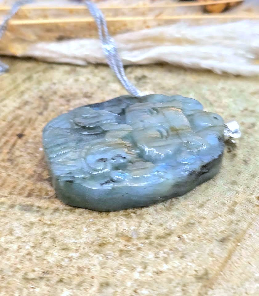 A carved jadeite pendant necklace featuring a deity, suspended from a silver-colored chain, displayed on a textured tan surface.