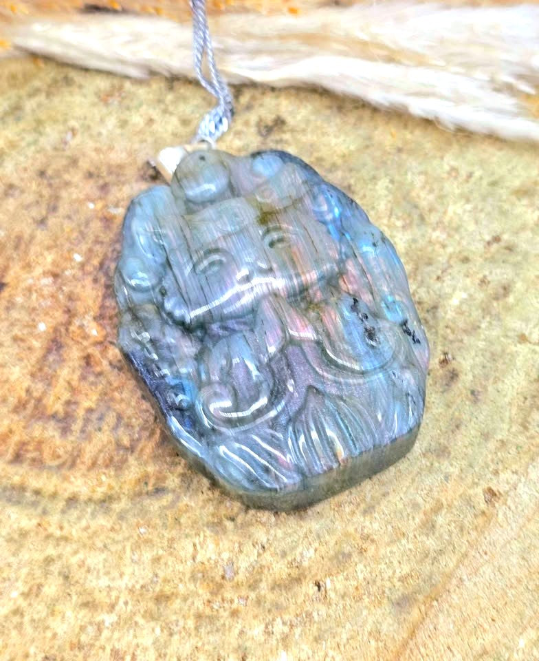 A carved labradorite pendant necklace featuring a stylized figure, resting on a textured sandy surface with a piece of driftwood in the background.