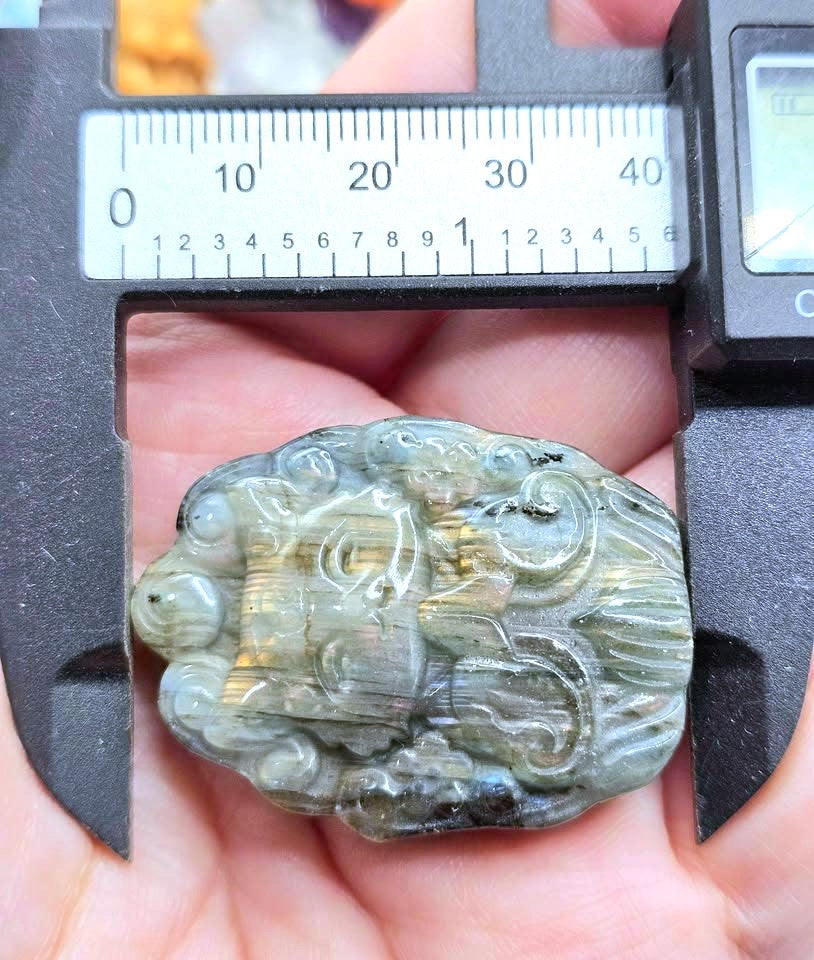 A close-up of a carved stone pendant or charm held in a hand under a digital caliper to show its size. The item appears to be a polished decorative piece with an abstract or animal-like carved form, in muted gray-green tones with some warm golden-brown highlights.