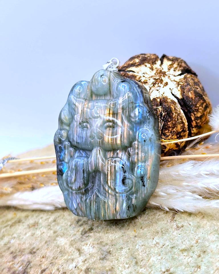 A carved blue stone pendant with a small silver loop at the top, displayed on a textured surface with a decorative shell and white feathers in the background.
