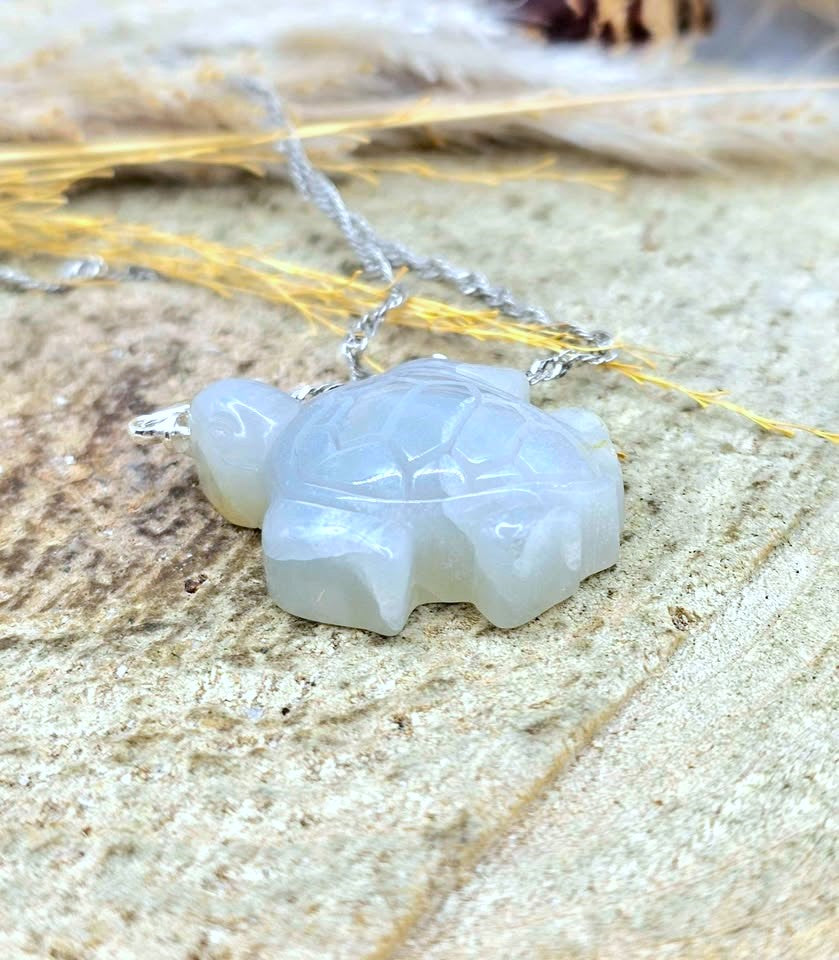 A carved turtle pendant necklace in a milky white translucent stone, suspended from a silver chain, resting on a textured stone surface with dried grass.