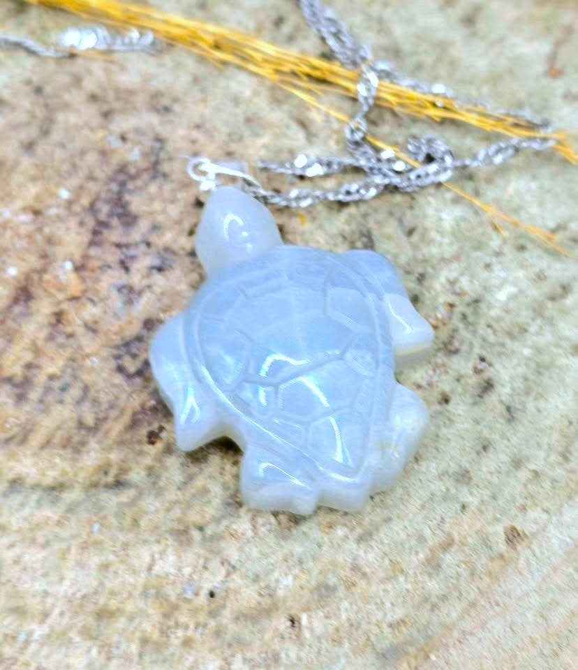 A carved turtle pendant necklace made of milky white jade, hanging from a silver chain on a textured stone surface.