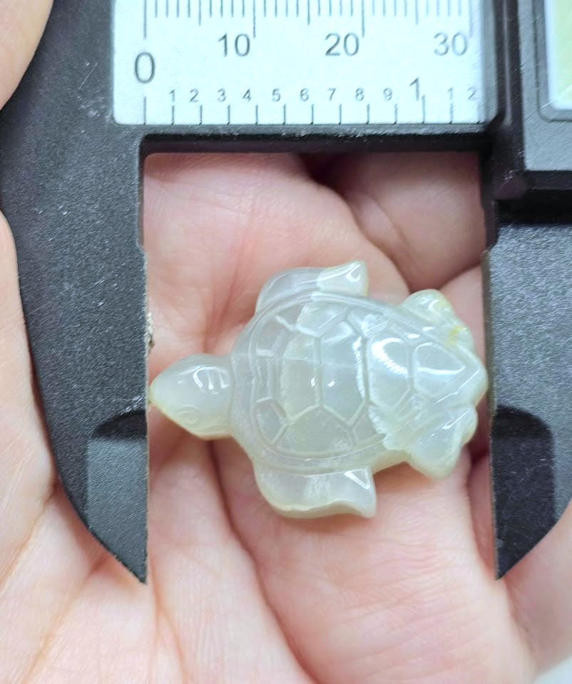 A small carved turtle figurine made of translucent pale green stone, held in a person's hand under a black caliper with a ruler scale visible at the top.