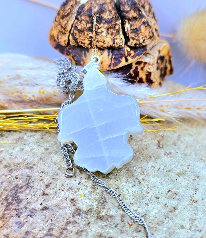 A silver-toned necklace with a translucent white, geometric, star-like pendant, displayed on a textured stone surface with dried grass and a decorative object in the background.