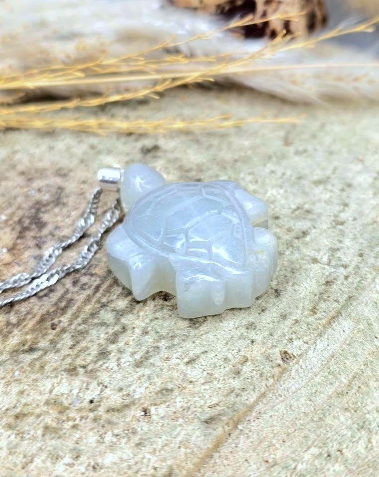 A carved turtle pendant necklace made of milky white stone, resting on a textured sandy surface with a silver chain partially visible.