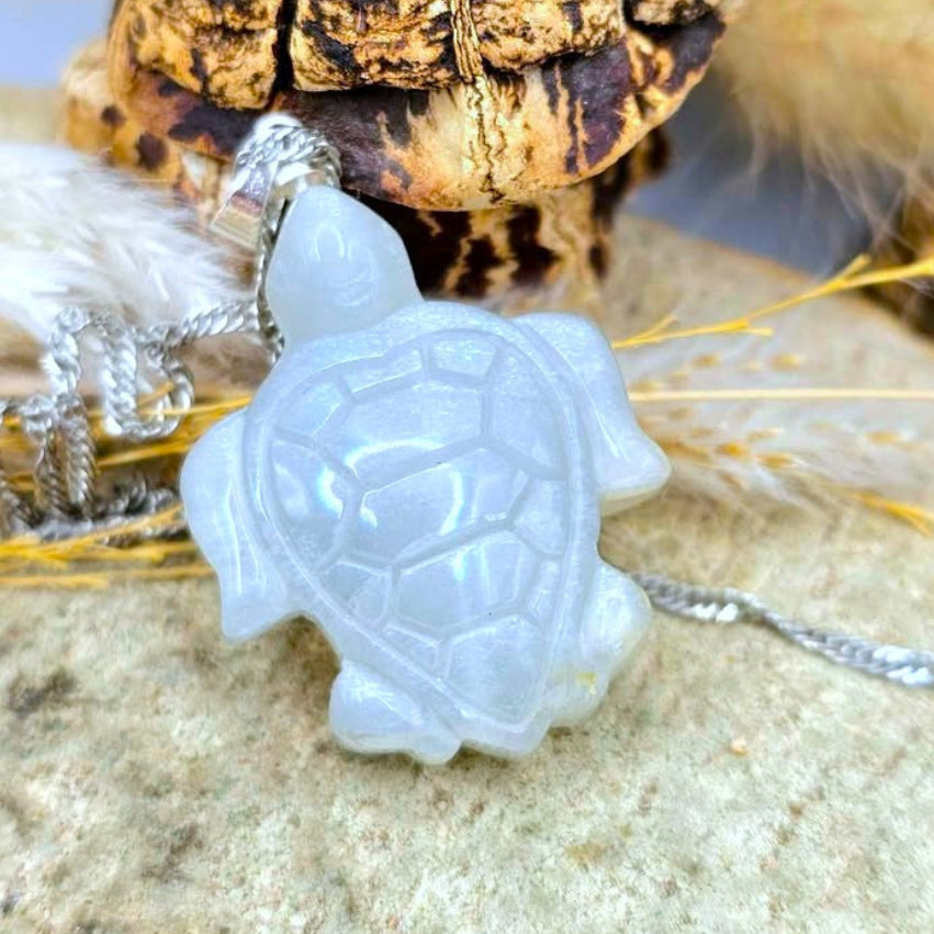 A carved sea turtle pendant made of pale translucent stone, hanging from a silver chain, styled on a rock with dried grass and a small turtle shell in the background.