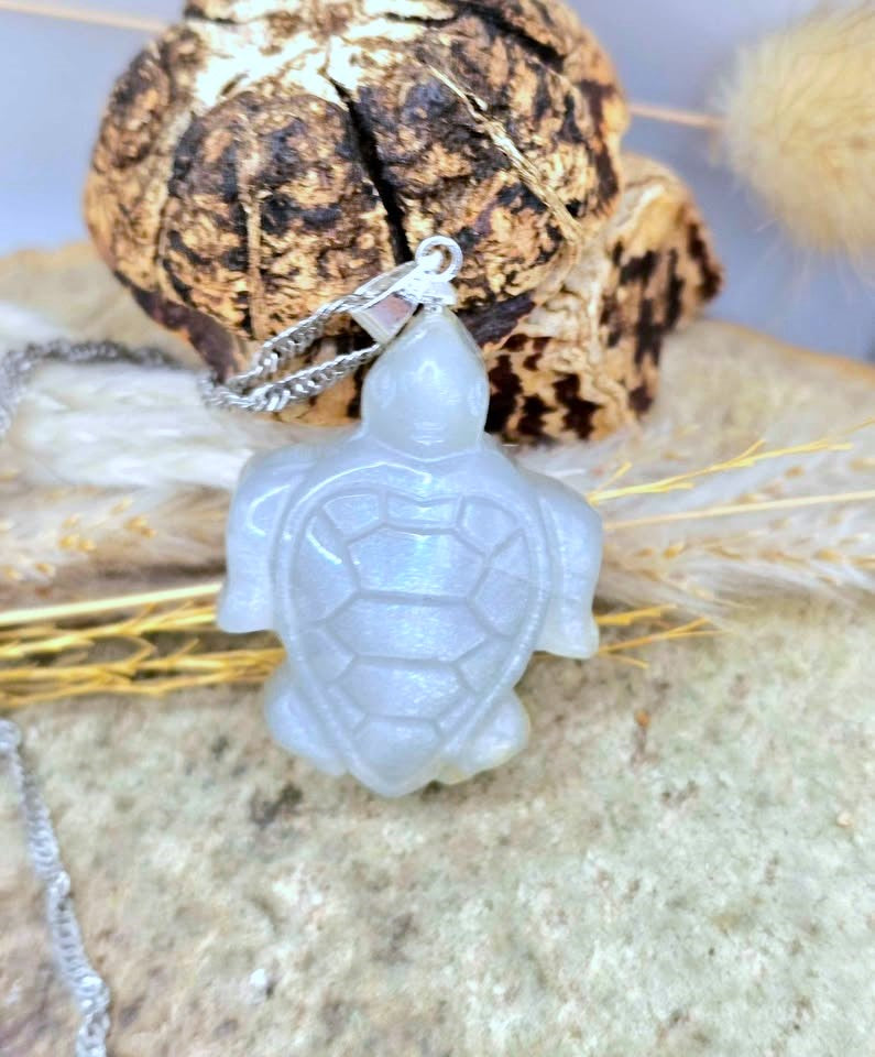 A carved turtle pendant necklace made of milky translucent white stone, hanging from a silver-toned chain, displayed on a textured stone surface with dried grass and bark in the background.