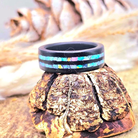 A black shungite ring with a colorful inlay of opal stone , displayed on a textured tree-bark stand with a pine cone and dried grass in the background.-My Aura Gemstones