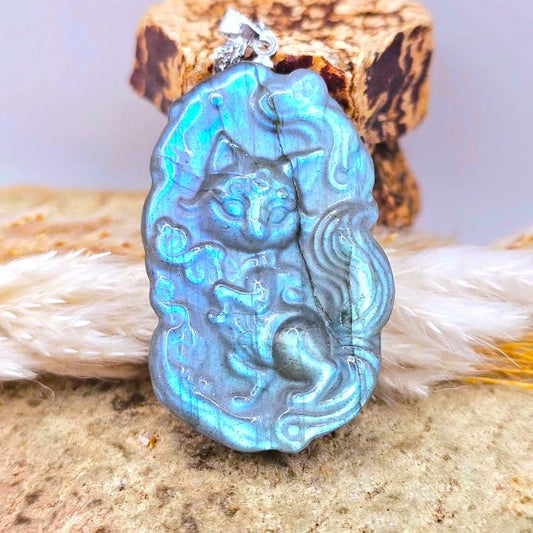A carved pendant featuring labradorite cat god with swirling blue, teal, and gold marbled detailing, resting on natural dried grasses and wood against a soft lavender background-My Aura Gemstones