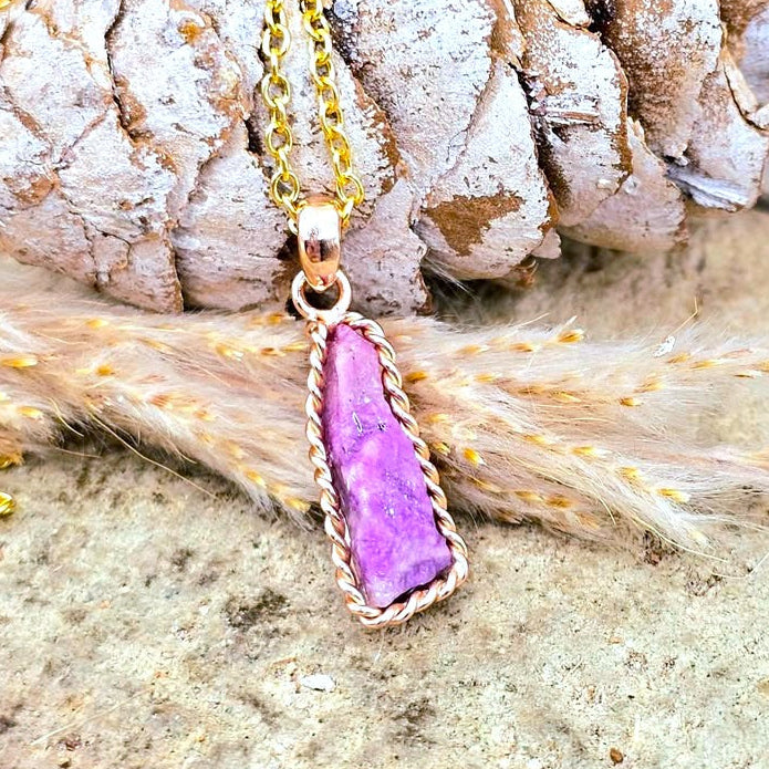 A gold chain necklace with a rectangular purple sugilite stone pendant in a twisted gold setting, styled on a textured stone surface with a pine cone and dried grass.-My Aura Gemstone
