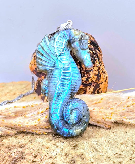 A labradorite carved sea horse-shaped pendant necklace with a metallic blue flash, resting on a piece of driftwood over sand against a soft gray background-My Aura Gemstones