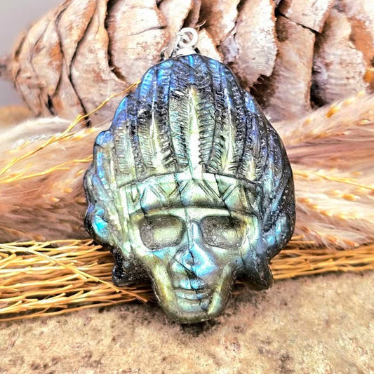 A skull-shaped pendant with a feathered headdress, featuring a blue-green iridescent finish and dark accents, displayed on a textured surface with dried grass and a pinecone in the background.