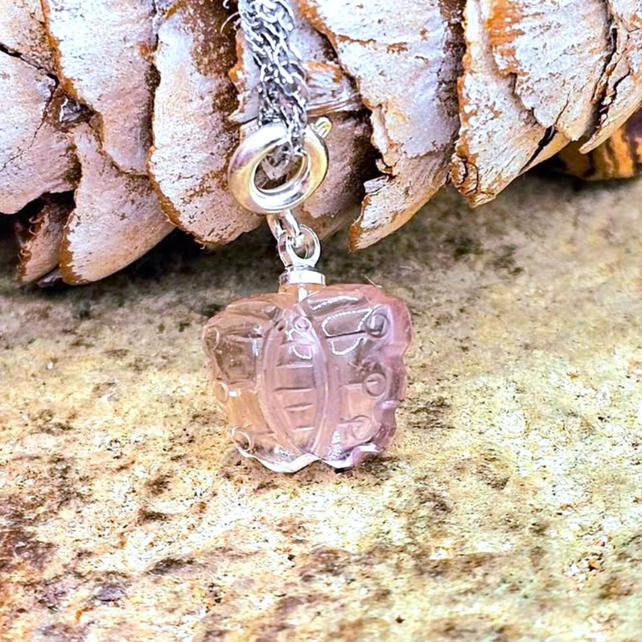 A silver chain necklace with a natural amethyst carved butterfly pendant resting against a textured rock and pinecone background.-My Aura Gemstones