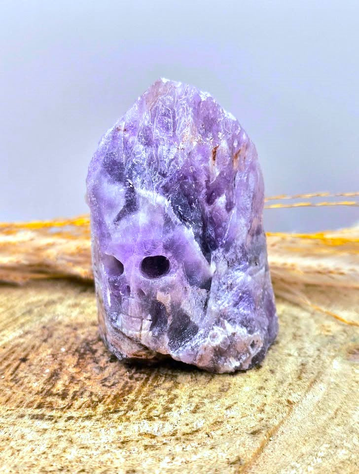 A carved natural Chevron Amethyst crystal skull figurine resting on a textured sandy surface with dried grass in the background.- My Aura Gemstones
