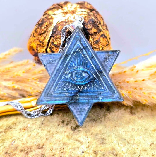 A labradorite blue flash all seeing eye pyramids pendant necklace featuring a Star of David and Eye of Providence design, resting on a textured stone with dried grass and a pinecone.-My Aura Gemstones