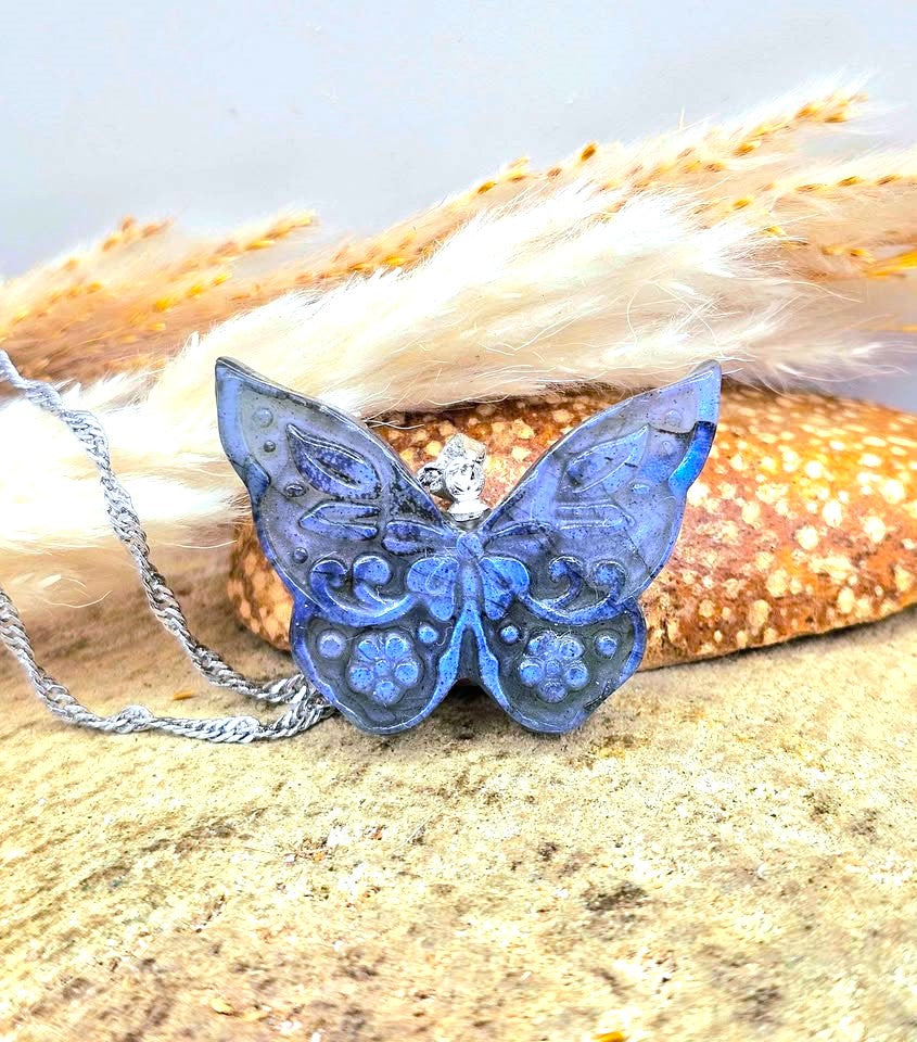 A blue butterfly-shaped pendant necklace with silver filigree detailing and a silver chain, displayed on a sandy surface with dried grass and a starfish prop.-My Aura Gemstones