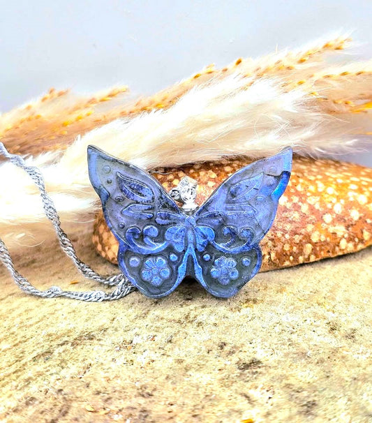 A blue butterfly-shaped pendant necklace with silver filigree detailing and a silver chain, displayed on a sandy surface with dried grass and a starfish prop.-My Aura Gemstones