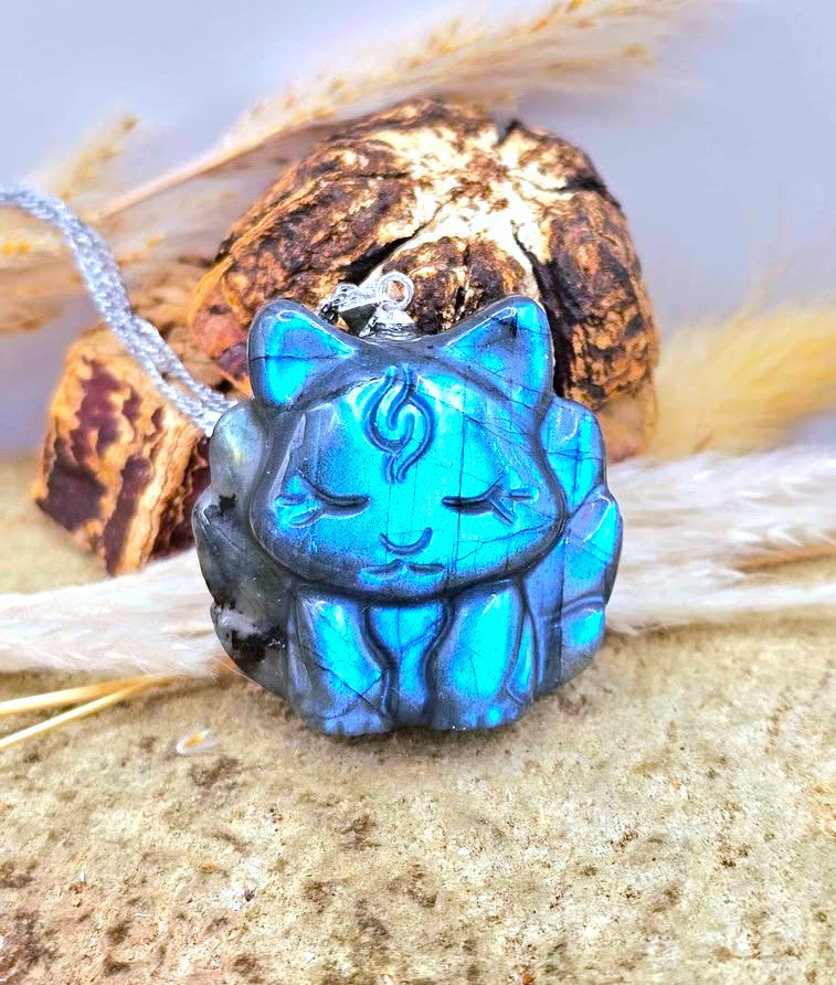 Labradorite baby nine tailed fox t- pendant with a vivid blue iridescent finish and dark gray edging, hanging from a silver chain and styled on sand with dried grass and a rock in the background.- My Aura Gemstones