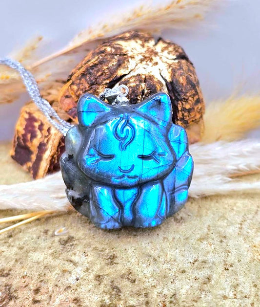 Labradorite baby nine tailed fox t- pendant with a vivid blue iridescent finish and dark gray edging, hanging from a silver chain and styled on sand with dried grass and a rock in the background.- My Aura Gemstones