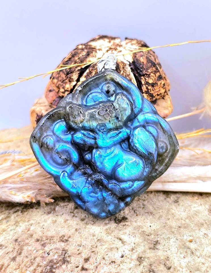 A carved blue labradorite pendant featuring a detailed sleeping baby fox design, displayed on a textured stone surface with dried grass and a small decorative object behind it.
