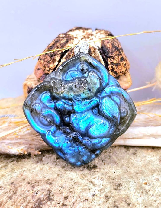 A carved blue labradorite pendant featuring a detailed sleeping baby fox design, displayed on a textured stone surface with dried grass and a small decorative object behind it.