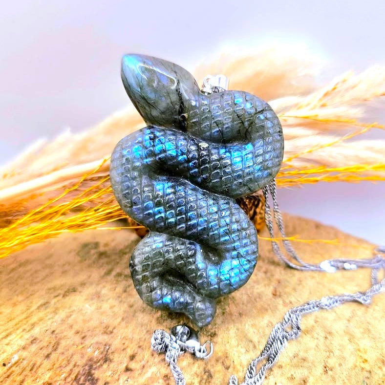 A coiled snake pendant necklace made of iridescent labradorite with a silver chain, displayed on a textured stone surface with dried grass.-My Aura Gemstones