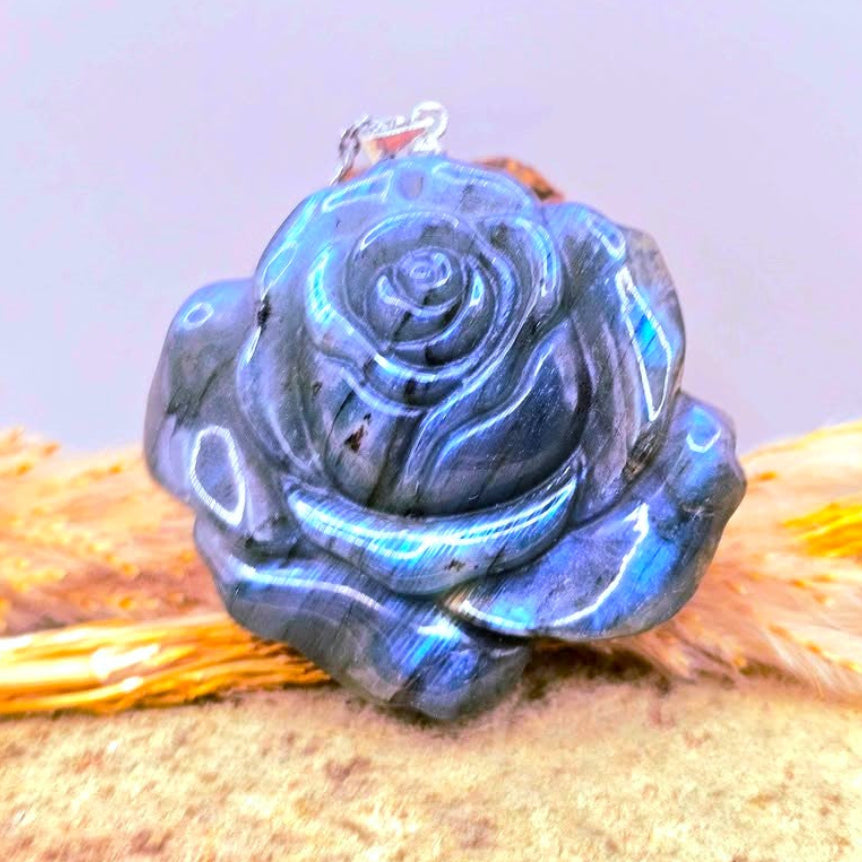 A labradorite carved blue flashy rose pendant with a silver bail, resting on a textured sandy surface with dried grass -My Aura Gemstones