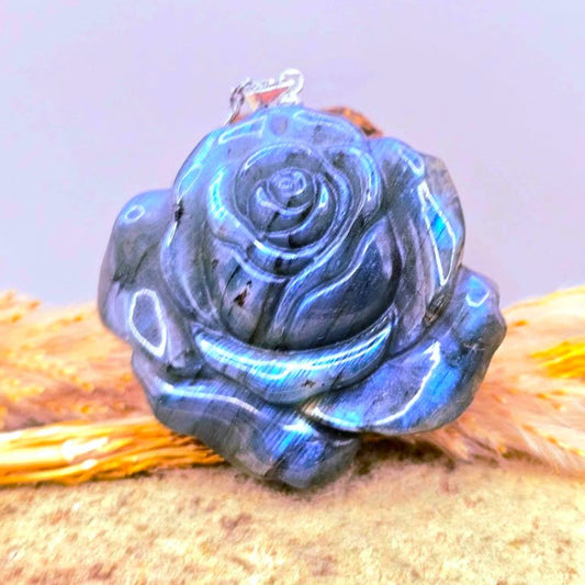 A labradorite carved blue flashy rose pendant with a silver bail, resting on a textured sandy surface with dried grass -My Aura Gemstones