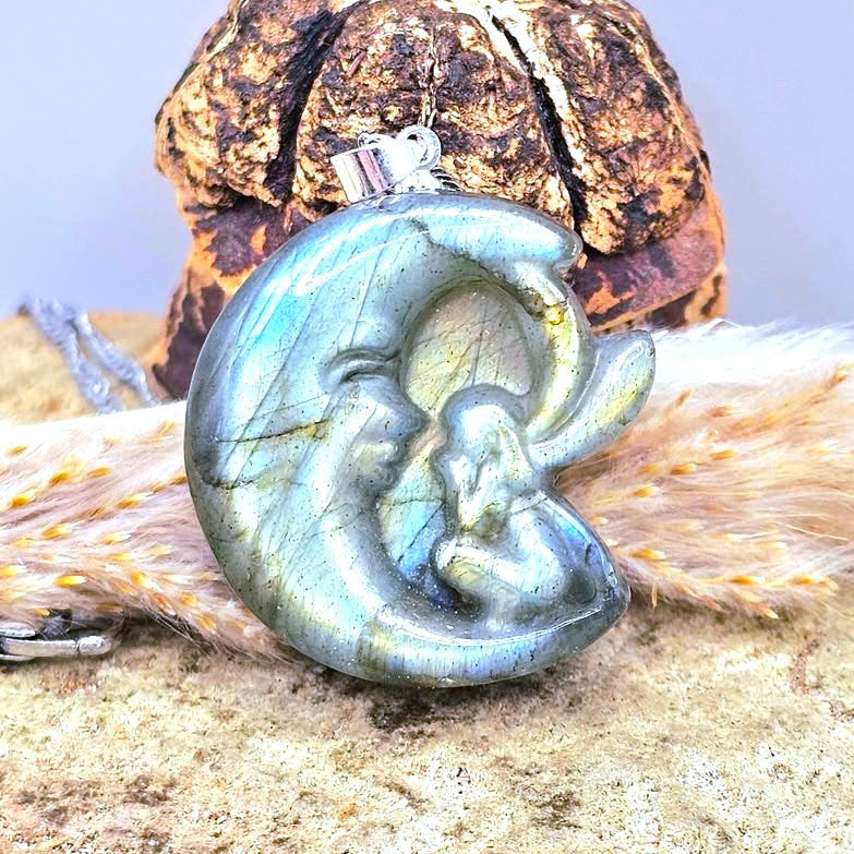 A flashy labradorite pendant shaped like a crescent moon with a butterfly fairy, resting on a textured surface with a dried mushroom and fur in the background.-My Aura Gemstones