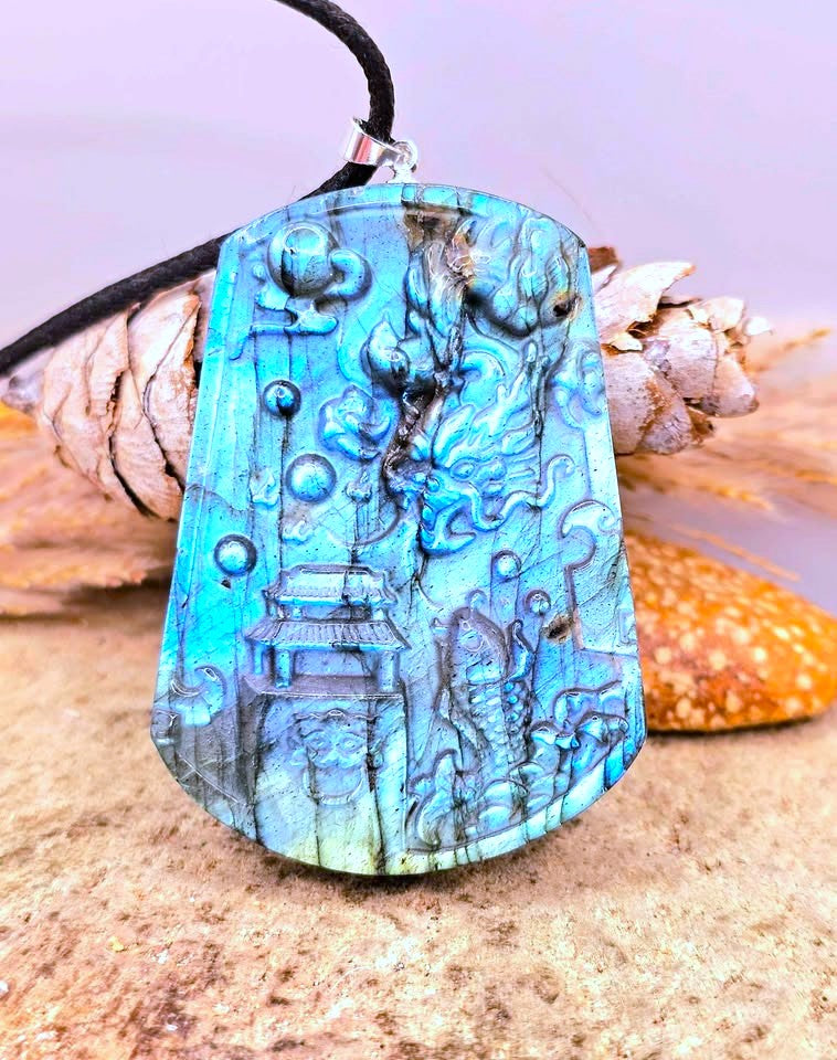 A carved blue labradorite pendant with craving of a flying dragon to a palace necklace with a black cord, displayed on a sandy surface with dried grass and a seashell.-My Aura Gemstones