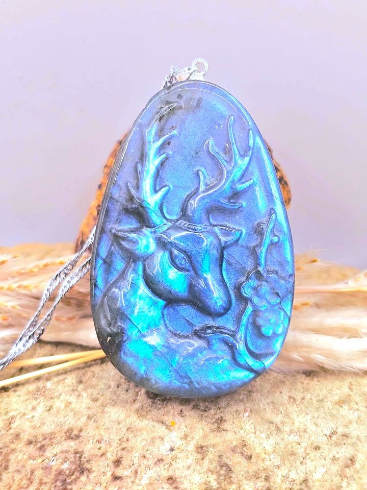 A teardrop-shaped labradorite pendant featuring a raised deer head design, resting on a textured stone surface with dried grass and a silver chain.-My Aura Gemstones
