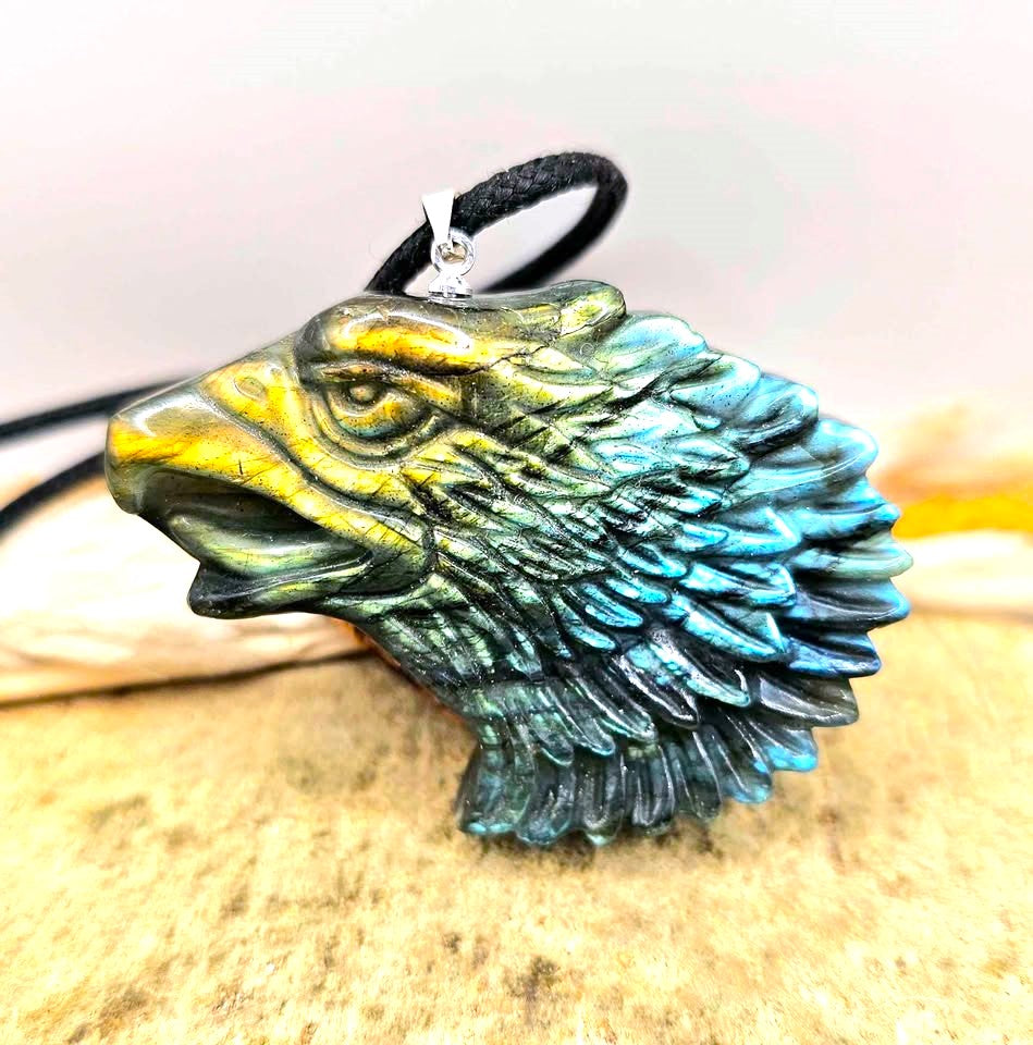 A carved eagle head pendant with a black cord, featuring a metallic gold beak and forehead transitioning to a blue-green patina across the feathers, resting on a textured stone surface-My Aura Gemstones
