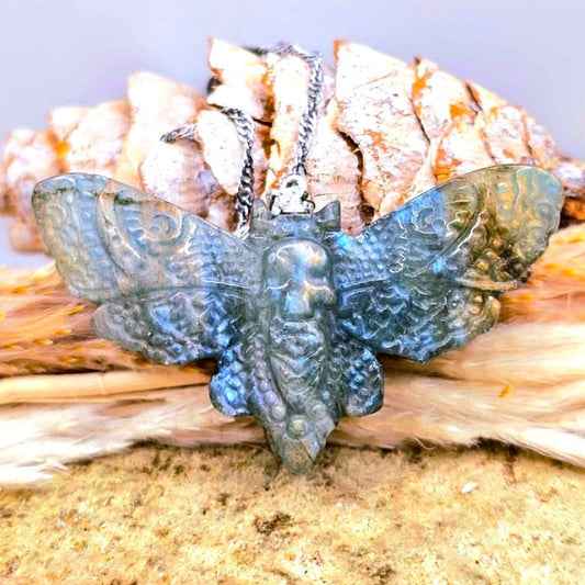 A detailed moth labradorite carved pendant necklace with a textured, metallic blue and green patina finish, resting on dried grass and wood against a soft gray background.